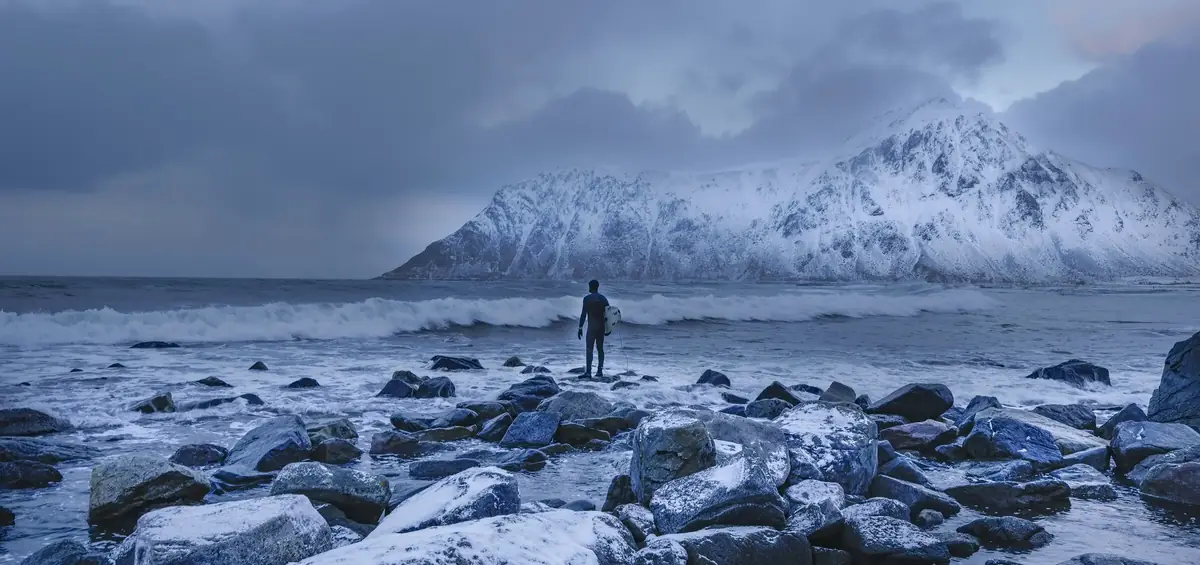 A surfer watching the waves under snowy mountains in Lofoten